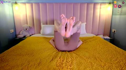 Media: Video of a woman lying on her back on a bed, legs bent, feet in the air, wearing a yellow blanket, in a cozy bedroom with pastel-colored walls, teddy bears, and warm lighting.