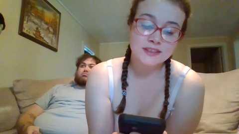 Media: Video of a young woman with glasses and braids, smiling, sitting on a beige sofa in a cozy living room. A man in a gray shirt rests behind her.