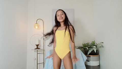 Media: Video of a young, slender Asian woman with long black hair, wearing a bright yellow one-piece swimsuit, standing in a minimalist room with a white wall, potted plant, and a lamp.