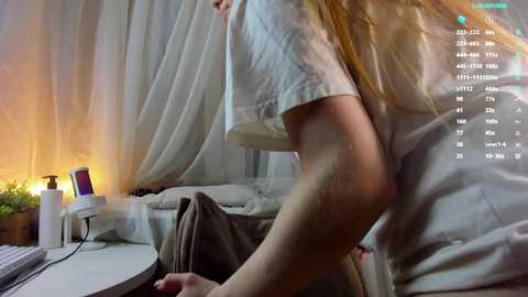 Media: Video of a woman with long blonde hair, wearing a white T-shirt, sitting on a bed with a gray blanket, in a dimly lit room with white curtains, digital screen overlay showing stats and chat.