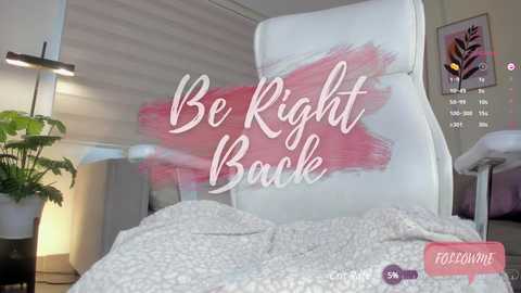 Media: Video of a modern, minimalist bedroom with a white leather recliner, pink abstract brushstroke wall art, and a potted plant. Text overlay reads \"Be Right Back\" in elegant white script.