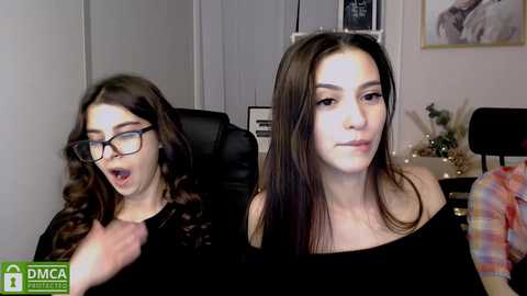 Media: Video of two young women with long dark hair, one wearing glasses, sitting in a dimly lit room. The background shows a black leather chair and a framed picture.