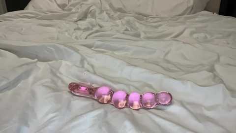 Media: A video of a pink, glass dildo with ridged texture on a rumpled white bedsheet in a dimly lit room. The dildo is centered and the sheets are disheveled.