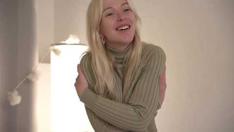 Media: Video of a smiling, fair-skinned woman with long blonde hair, wearing a green knitted sweater, standing in a softly lit room with a white wall and a cylindrical lamp.