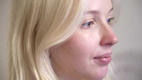 Media: Video of a close-up, side-profile view of a young Caucasian woman with fair skin, straight blonde hair, and freckles. She has a gentle, slightly closed-mouth smile. Background is a plain, neutral-colored wall.