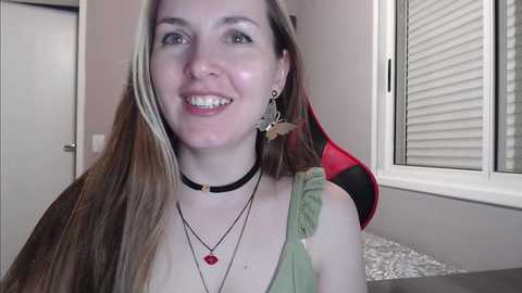 Media: Video of a smiling, fair-skinned woman with long, straight, blonde hair, wearing a black choker necklace, green top, and multiple layered necklaces with a red pendant. Background includes a window with closed blinds and a red gaming chair.