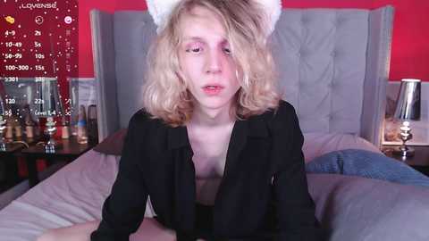 Media: Video of a young, pale-skinned, blonde woman with shoulder-length hair wearing a black top and white cat ears, lying on a bed with a gray headboard.