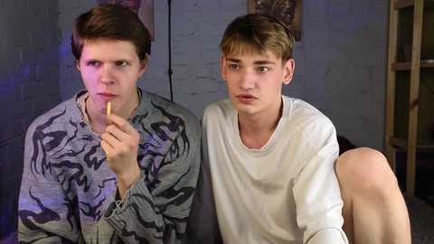 Media: Video of two young boys in a dimly-lit room, one holding a cigarette, both dressed casually in t-shirts, against a gray brick wall.