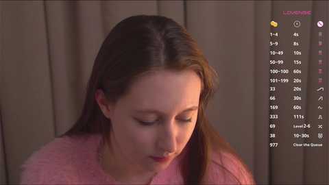 Media: Video of a young woman with light skin and long brown hair, wearing a pink fuzzy sweater, against a beige curtain background.