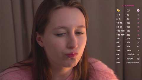 Media: Video of a young Caucasian woman with long brown hair, wearing a pink fuzzy coat, looking down with a pensive expression. Background shows a beige curtain.