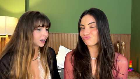 Media: Video of two women with long hair, one blonde and one brunette, smiling and making kissing faces, standing in a green-walled room with a wooden headboard and nightstand.