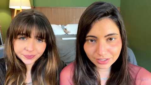 Media: Video of two young women with light skin, dark hair, and light makeup, smiling; one wears a black top, the other a red shirt. Background: green wall, bed, lamp, and neutral-toned furniture.