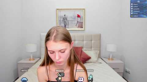 Media: Video of a young woman with straight, light brown hair, wearing a black top, standing in a minimalist bedroom with a beige upholstered headboard, framed artwork, and nightstands.