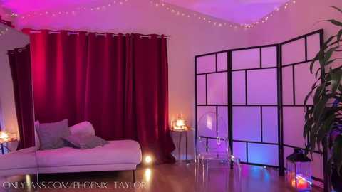 Media: Video of a cozy, dimly lit room with a white daybed, burgundy curtains, black folding screen, and string lights. Soft purple and pink hues illuminate the space.