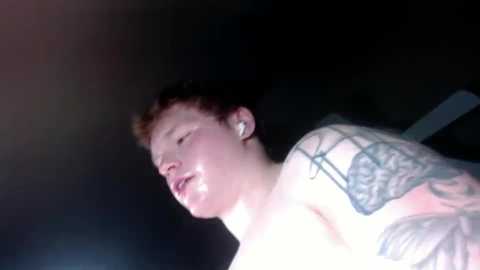 Media: Video of a shirtless man with short, curly brown hair, eyes closed, and a large black tattoo on his right shoulder, set against a dark, blurry background.