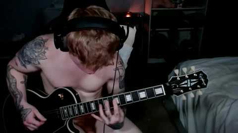 Media: Video of a shirtless, muscular man with tattoos, red hair, and large headphones, playing an electric guitar in a dimly lit bedroom with a bed and shelves in the background.