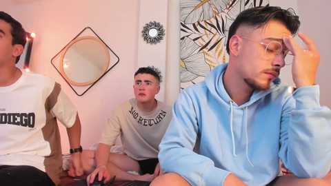 Media: Video: Three young men in casual clothes sit in a cozy room with a circular mirror, wall art, and string lights.