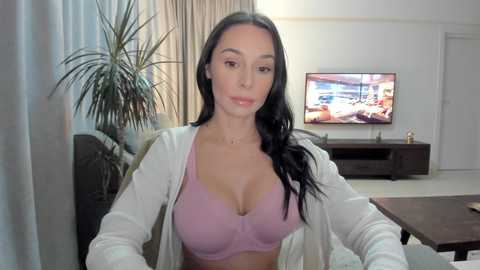 Media: Video of a young woman with long black hair, fair skin, and large breasts wearing a lavender bra, seated in a modern living room with a TV displaying an action scene.