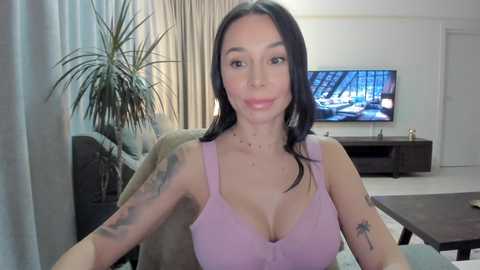 Media: Video of a smiling, tattooed woman with long black hair, wearing a low-cut lavender tank top, sitting in a living room with a flat-screen TV and potted plant.