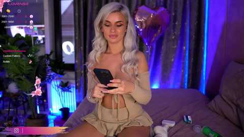 Media: Video of a blonde woman with wavy hair, wearing a beige crop top and matching shorts, sitting on a bed with a gaming setup in the background.