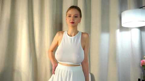 Media: Video of a fair-skinned, blonde woman with large breasts wearing a white, sleeveless crop top and high-waisted skirt, standing in front of light-colored curtains and a modern lamp.