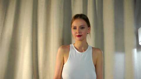 Media: Video of a fair-skinned woman with light brown hair in a bun, wearing a white sleeveless top, standing against beige curtains.