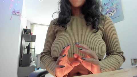 Media: Video of a woman with medium-dark skin, long wavy black hair, and a beige ribbed sweater, lifting her shirt to reveal her large breasts. She has black nail polish. Background includes a white wall, a shelf, and a poster.