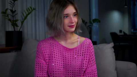 Media: Video of a young woman with short, light brown hair, wearing a pink, crochet-patterned sweater, sitting on a gray couch in a dimly-lit living room with potted plants.