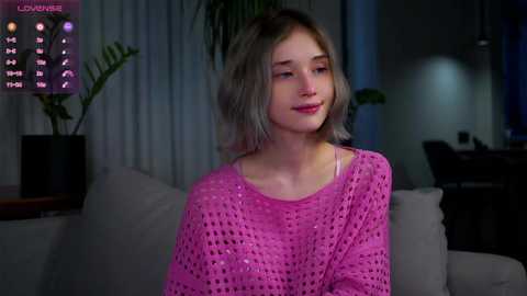 Media: A young woman with shoulder-length blonde hair sits on a couch, wearing a pink, knitted sweater, in a dimly lit room with a calendar and green potted plants.