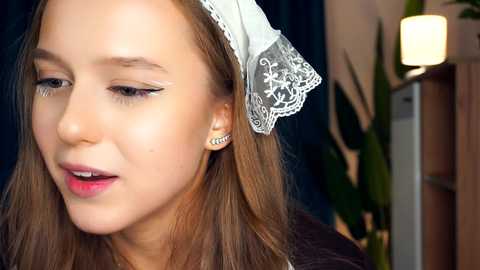 Media: Video of a young woman with fair skin, straight light brown hair, and delicate makeup. She wears a white lace headband and small silver stud earrings, standing indoors with a blurred background.