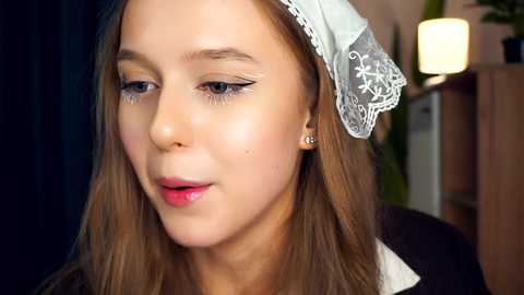 Media: Video of a young Caucasian woman with fair skin, light brown hair, wearing a white lace headband, light makeup, and a black dress. Background shows blurred indoor setting with a lamp and plant.