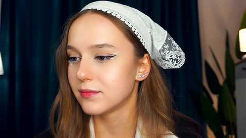 Media: Video of a young, fair-skinned woman with light brown hair, wearing a white lace headscarf and silver stud earrings, with dark eyeliner, set against a dark blue curtain backdrop.