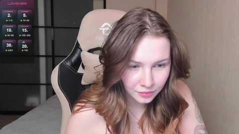 Media: Video of a fair-skinned, brunette woman with long, wavy hair, wearing a black bra, sitting on a beige gaming chair in a modern, minimalist room.