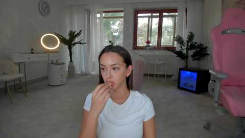 Media: Video of a young woman with long brown hair, wearing a white t-shirt, biting her finger, standing in a minimalist, white-walled room with a large window, desk, and pink gaming chair.