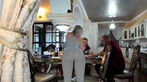 Media: Video of a lavish, ornate dining room with chandeliers, elegant curtains, and four people; one woman in gray workout attire center, another in a black top and glasses, and two men in casual clothes.