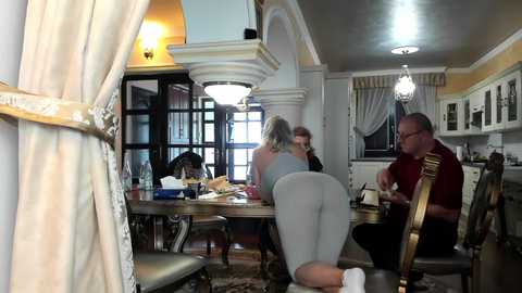 Media: Video of a spacious, ornate dining room with three people. A woman in gray leggings bends over a table, a man in glasses sits nearby, and a woman with blonde hair and glasses stands.