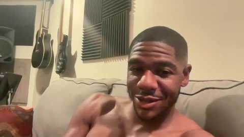 Media: Video of a shirtless, muscular Black man with a shaved head, sitting on a beige couch, smiling. Wall-mounted guitars and a dark acoustic guitar hang in the background.