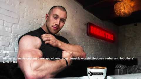 Media: Video of a muscular bald man with a shaved head, wearing a black sleeveless shirt, flexing his arm, sitting at a cafe table with a white cup and saucer. Background shows a brick wall and a red neon sign.