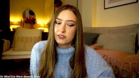 Media: Video of a young woman with long, straight brown hair, wearing a light blue sweater, sitting indoors near a bed with beige pillows and a pink blanket.