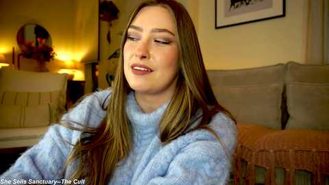 Media: Video of a young woman with long, straight brown hair, wearing a light blue sweater, sitting indoors. Background features a beige couch, framed artwork, and a warm, cozy ambiance.