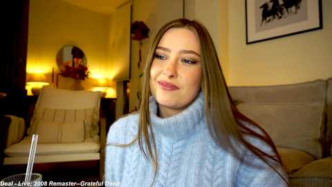 Media: Video of a smiling, light-skinned woman with long, straight brown hair, wearing a light blue, high-necked sweater, seated in a cozy living room with yellow walls and framed artwork.