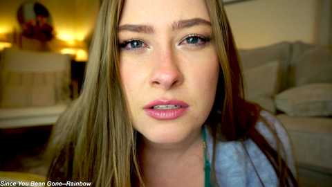 Media: Video of a young woman with long blonde hair, light skin, and blue eyes, wearing a denim jacket, sitting in a living room with beige furniture.