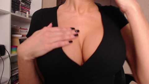 Media: Video of a woman with large breasts in a black V-neck shirt, revealing cleavage. She has fair skin and black-painted nails. Background shows white shelves with books and a rainbow-colored blanket.