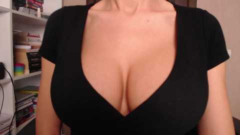 Media: Video of a woman with large breasts, wearing a plunging black top, in a home office setting with shelves filled with books and colorful folders.