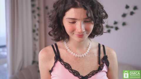 Media: Video of a young woman with curly dark hair, wearing pink camisole with black lace trim and pearl necklace, in a softly lit room with greenery on the wall.