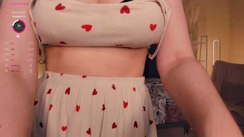 Media: Video of a fair-skinned woman in a beige ribbed crop top with red heart patterns and matching high-waisted skirt. Background shows a cozy room with a bed and a nightstand.