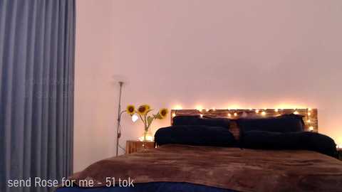 Media: Video of a modern bedroom with a white wall, a dark brown bed with a wooden headboard, two sunflower vases, and a blue curtain on the left.