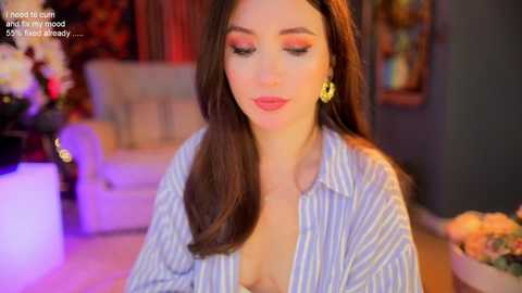 Media: Video of a fair-skinned woman with long brown hair, wearing a blue and white striped button-down shirt, with a low neckline revealing cleavage. She has bold red lipstick and is smiling softly. Background features a blurred, cozy living room with warm lighting.