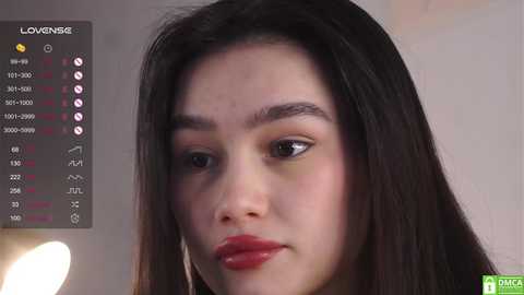 Media: A close-up video of a young Asian woman with long black hair, light skin, and red lipstick, looking pensive. The background shows a monitor displaying a weather forecast.