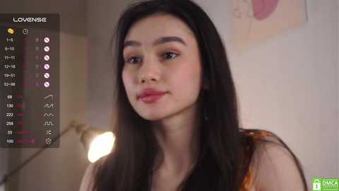 Media: A video of a young Asian woman with long black hair, fair skin, and light makeup, wearing a black sleeveless top. She's indoors with a blurred background featuring a soft light and a poster.